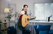 © Parichat - Young woman is playing an acoustic guitar in her living room, wearing headphones and making the rock sign with her hand while smiling