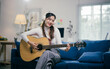 © Parichat - Young woman playing acoustic guitar in her cozy living room, fully immersed in music, showcasing talent and creativity