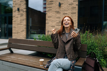 Wall Mural - Cheerful redhead woman receiving unexpected good news in social media app using smartphone sitting on urban bench on summer day. Happy female celebrating success with joy and positive vibes.