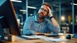 © Sumet - A thoughtful man with a beard rests his head in his hand, contemplating while seated at a desk with a computer in a dimly lit office environment.