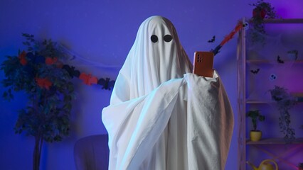 Funny halloween ghost in white sheet with black eyes in dark room talking on smartphone by video call, positive expression. Happy halloween spooky concept.