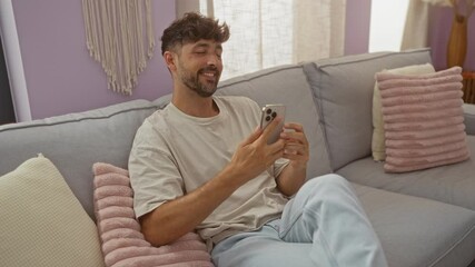 Wall Mural - Young man relaxing on a gray sofa at home using a smartphone in a cozy living room