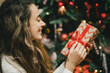 © polinaloves - Young woman holding Christmas present with a red ribbon, standing near Christmas tree.