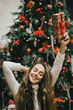 © polinaloves - Young woman holding Christmas present with a red ribbon, standing near Christmas tree.