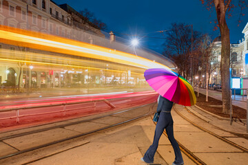 Wall Mural - A beautiful young girl with a multicolored umbrella crosses the street - Tram moving on a street at twilight blue hour - Vienna, Austria