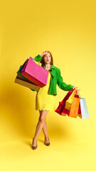 Giant discounts. Full-length image of beautiful young woman in stylish clothes standing with multicolored shopping bags against yellow background. Shopping, sales, Black Friday, Cyber Monday concept