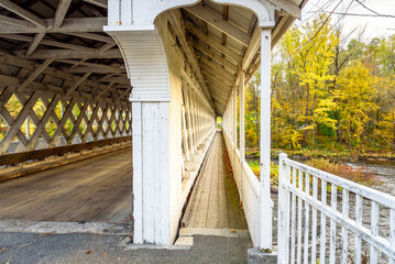 Wall Mural - Empty walkway along the side of a wooden covered bridge spanning a river with forested banks at the peak of fall foliage