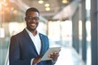 © VK Studio - A confident young man in a suit and glasses, smiling while using a tablet in a modern office space, exuding professionalism.
