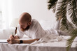 © Andrii Lysenko - Bearded young man laying on bed and writing in diary at home.