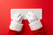© ActionGP - Santa Claus hands in gloves typing on a white keyboard against a red background symbolizing holiday preparations and Christmas communication