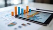© BerkahStock - A tablet displaying colorful graphs and charts alongside printed reports on a table.