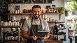 © h4kunA - Happy barista in apron standing behind the counter with a tablet in his hands, smiling.