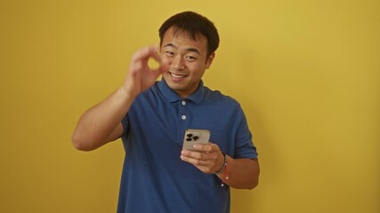 Sticker - Young chinese man using smartphone with smiling and friendly ok sign gesture over isolated yellow background, displaying confident positive expression and excellent symbol with finger