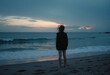 © Derek Brumby - A solitary figure stands on a beach, gazing at a serene sunset over the ocean, symbolizing solitude, reflection, and the beauty of nature and stillness.