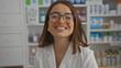 © Krakenimages.com - Smiling brunette woman with glasses and white coat standing in a pharmacy, showcasing a friendly atmosphere among shelves filled with pharmacy products.