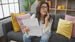 © Krakenimages.com - A concerned hispanic woman talks on the phone in her living room holding documents.