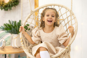 Cute happy laughing little girl in dress sits in a wicker hanging chair and ride on Christmas morning at home Happy winter holidays. High quality photo