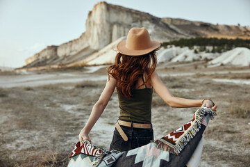 Wall Mural - Serene woman in widebrimmed hat poses against desert backdrop with colorful blanket and majestic mountains