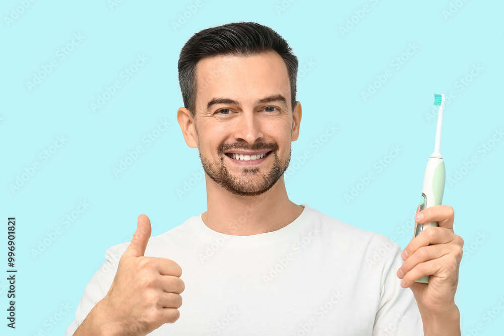 Handsome man with electric toothbrush showing thumb-up on blue background