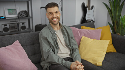 Wall Mural - A cheerful young man with a beard comfortably seated in a modern living room featuring colorful pillows and home decor.