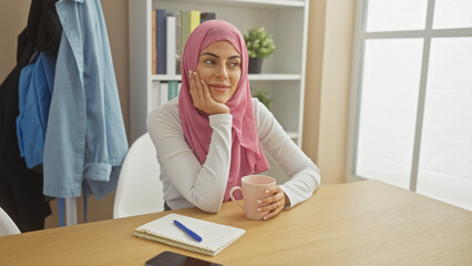 Wall Mural - Smiling woman wearing hijab holding mug in modern home office