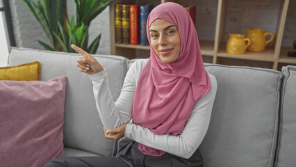 Poster - A smiling young woman in a pink hijab sitting on a gray couch in a modern living room, gesturing to the side.