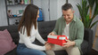 © Krakenimages.com - A man and woman share a joyful moment as he receives a red giftbox from her in their cozy living room.
