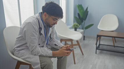 Wall Mural - A young bearded man in medical attire using a smartphone in a modern clinic waiting room.