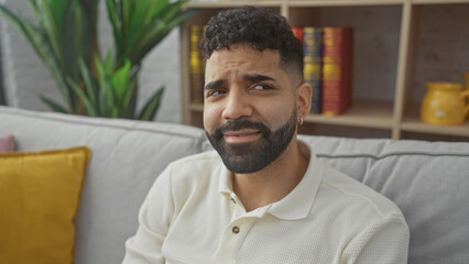 Sticker - A young handsome hispanic man with a beard poses in a cozy home interior with a plant and bookshelf.