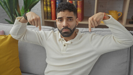 Sticker - A young hispanic man with a beard gestures downward while sitting on a gray couch indoors, conveying a casual yet expressive atmosphere.