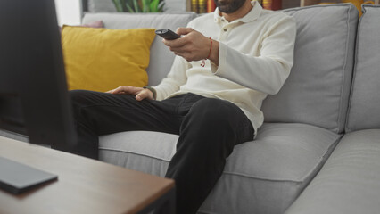 Sticker - A young man with a beard sitting on a gray couch, holding a remote and watching television at home.