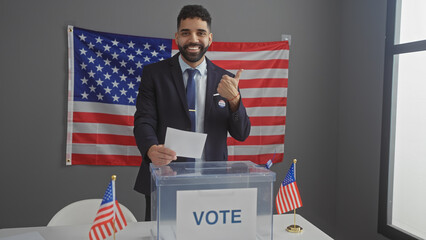 A smiling young hispanic man with a beard giving thumbs up after voting in an american indoor electoral setting with flags.