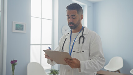 Wall Mural - Handsome hispanic man, wearing a lab coat and stethoscope, takes notes in a bright clinic room.