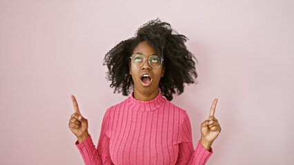 Wall Mural - African woman in glasses expressing surprise indoors with a pink background, pointing upwards.