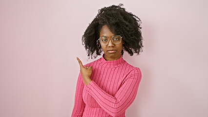 Wall Mural - African american woman in glasses pointing sideways in a pink sweater against a plain background.