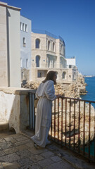 Wall Mural - A beautiful young hispanic woman in white clothing enjoys the scenic coastal view from a viewpoint in polignano a mare, puglia, italy, with historic buildings and clear blue skies in the background.