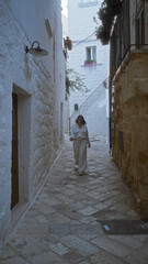 Wall Mural - Young hispanic woman walking along the beautiful narrow streets of polignano a mare, puglia, italy, surrounded by charming white buildings.