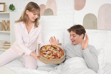 Wall Mural - Young woman holding cardboard box with tasty pizza and her surprised boyfriend sitting on bed in bedroom