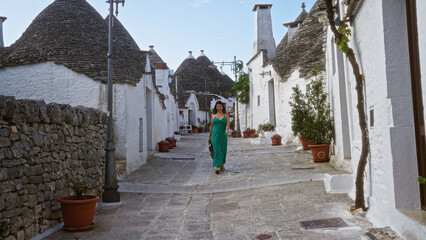 Wall Mural - Young hispanic woman in a green dress walking through the picturesque stone streets of alberobello, italy, surrounded by traditional trulli houses under a clear blue sky.