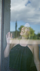 Poster - A young hispanic woman smiling indoors, her hand touching a glass window, creating a reflective effect with a scenic outdoor background.