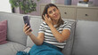 © Krakenimages.com - A young hispanic woman is sitting on a couch in her living room, holding a mobile phone and adjusting her hair, while wearing a striped shirt and covered with a blue blanket.