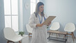 © Krakenimages.com - A young female doctor stands in a hospital waiting room holding a clipboard, surrounded by chairs, bright lighting, and a clock on the wall.