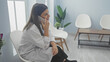 © Krakenimages.com - A young, attractive, hispanic, brunette woman in a lab coat talks on the phone in a clinic waiting room with modern white chairs and plants.