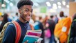 © Elmira - A high school graduate stands excitedly at a college fair booth, holding a stack of university brochures while interacting with other attendees in the background