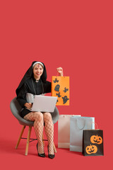 Sticker - Young woman dressed as nun for Halloween with laptop and shopping bags on red background