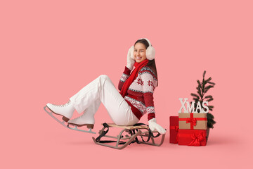 Sticker - Happy young woman in ice skates with gift boxes, Christmas tree and word XMAS on sledge against pink background