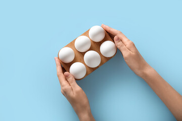 Poster - Female hands with wooden holder of raw chicken eggs on blue background