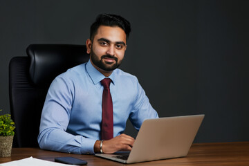 Canvas Print - Handsome young Indian businessman sitting at his desk in the office with a laptop