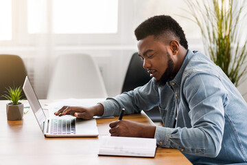 Wall Mural - African American employee using laptop and taking notes while working in office, side view with copy space