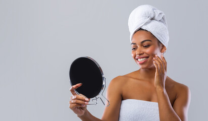 Wall Mural - Satisfied smiling black woman wrapped in towel checking her face in mirror after shower over grey background, skincare concept